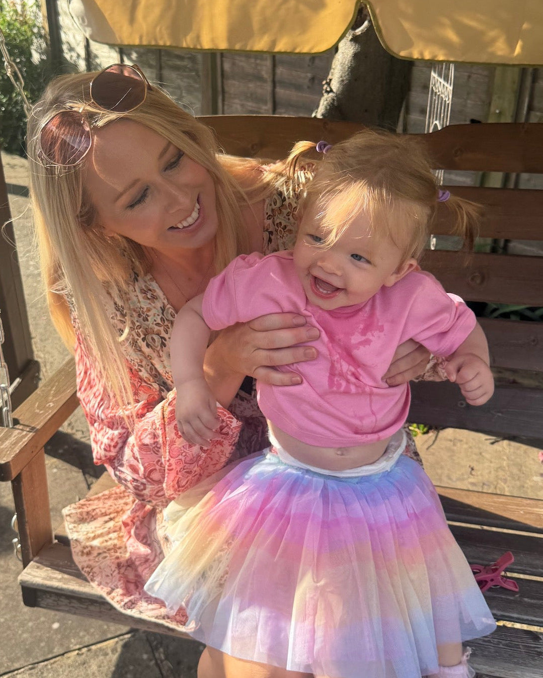 Happy mother holding her smiling toddler wearing a pink shirt and rainbow tutu while sitting together on a wooden bench outdoors